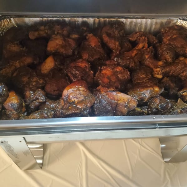 A catering tray filled with dark, saucy Jamaican Cafe Cuisine jerk chicken pieces.
