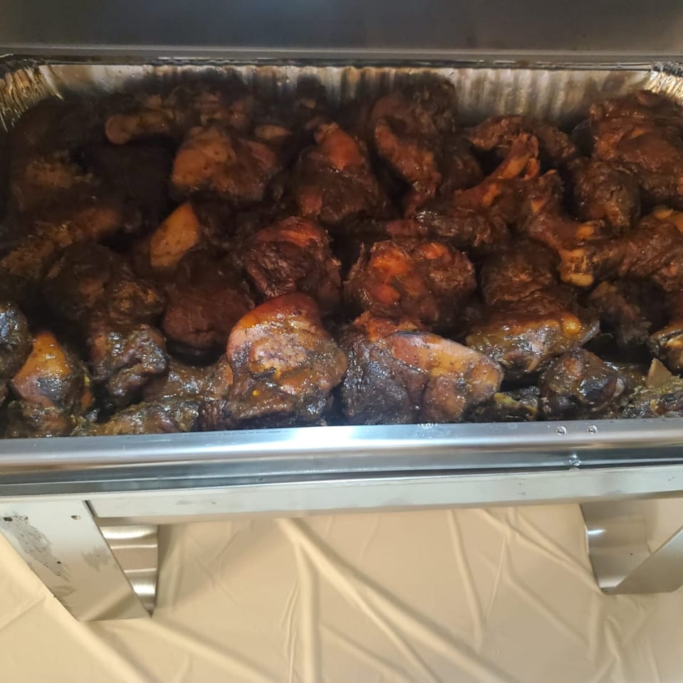 A catering tray filled with dark, saucy Jamaican Cafe Cuisine jerk chicken pieces.