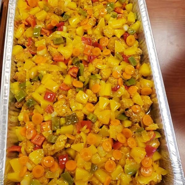 A large catering tray filled with Jamaican sweet and sour chicken, bell peppers, carrots, and pineapple.