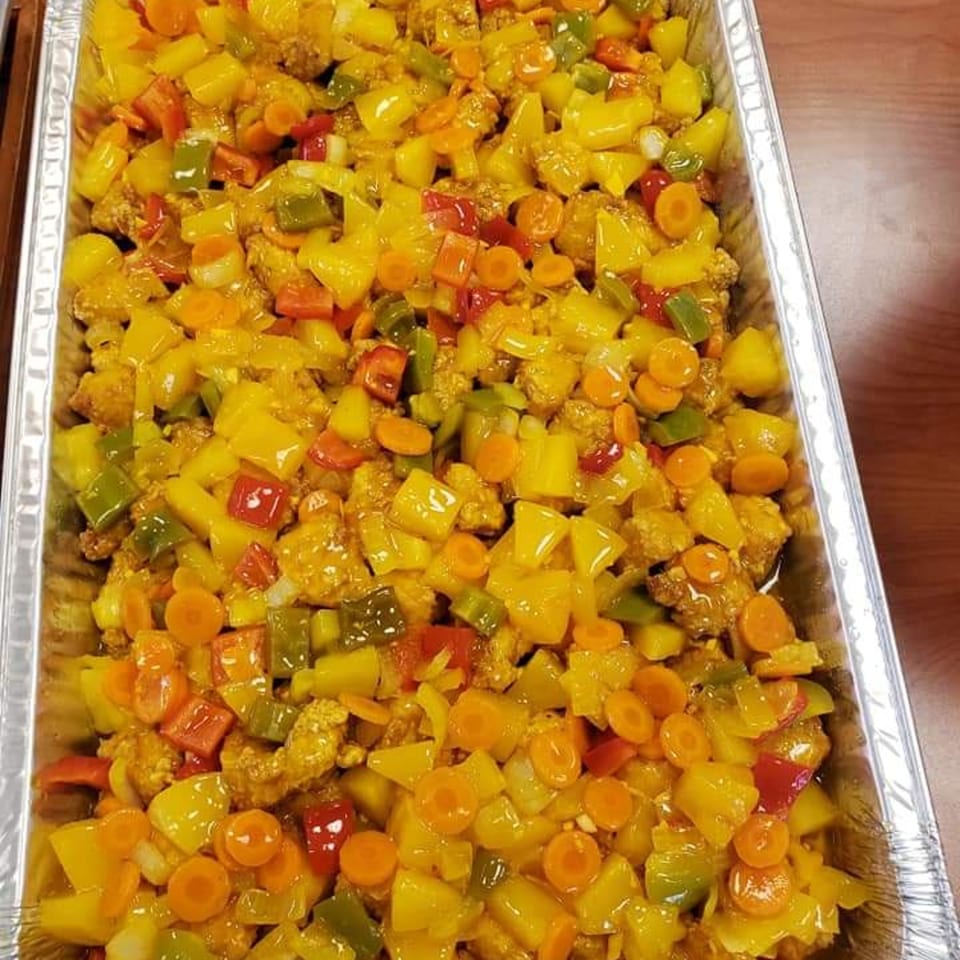 A large catering tray filled with Jamaican sweet and sour chicken, bell peppers, carrots, and pineapple.