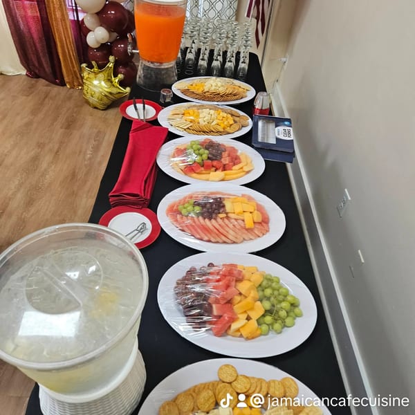A buffet table with fruit platters, cheese and crackers, and drink dispensers at Jamaican Cafe Cuisine.